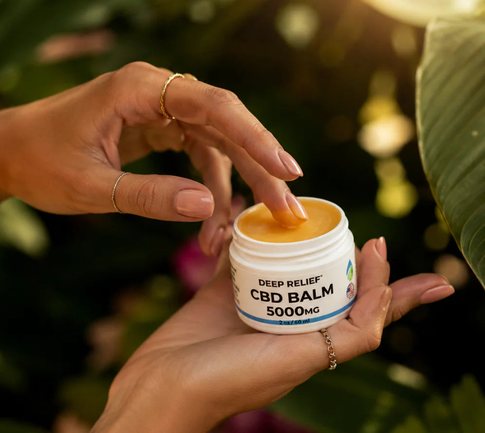 Hands holding an open Deep Relief CBD Balm jar, with a finger dipping into golden balm against a sunlit garden background.